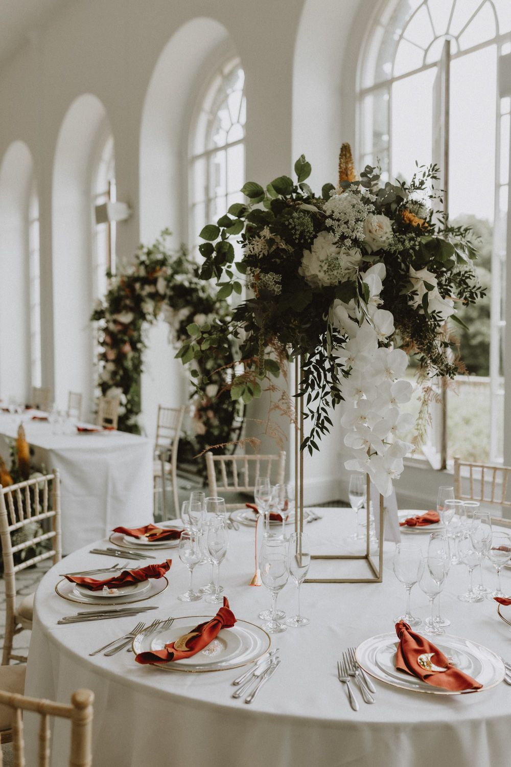 Elegant wedding reception table setting with tall floral centerpieces and antique white decor in a bright venue with arched windows.