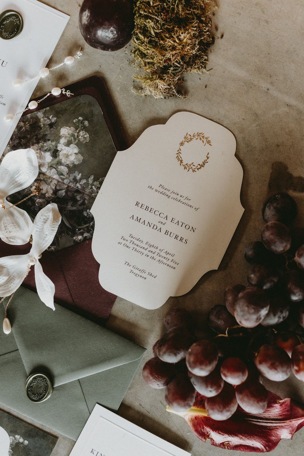 Wedding invitation with ornate design displayed among grapes and envelopes.