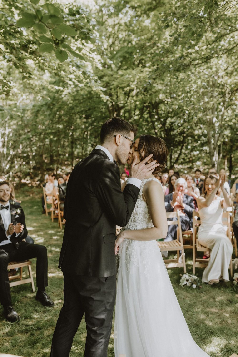 An outdoor wedding ceremony kiss surrounded by guests in a wooded garden setting.