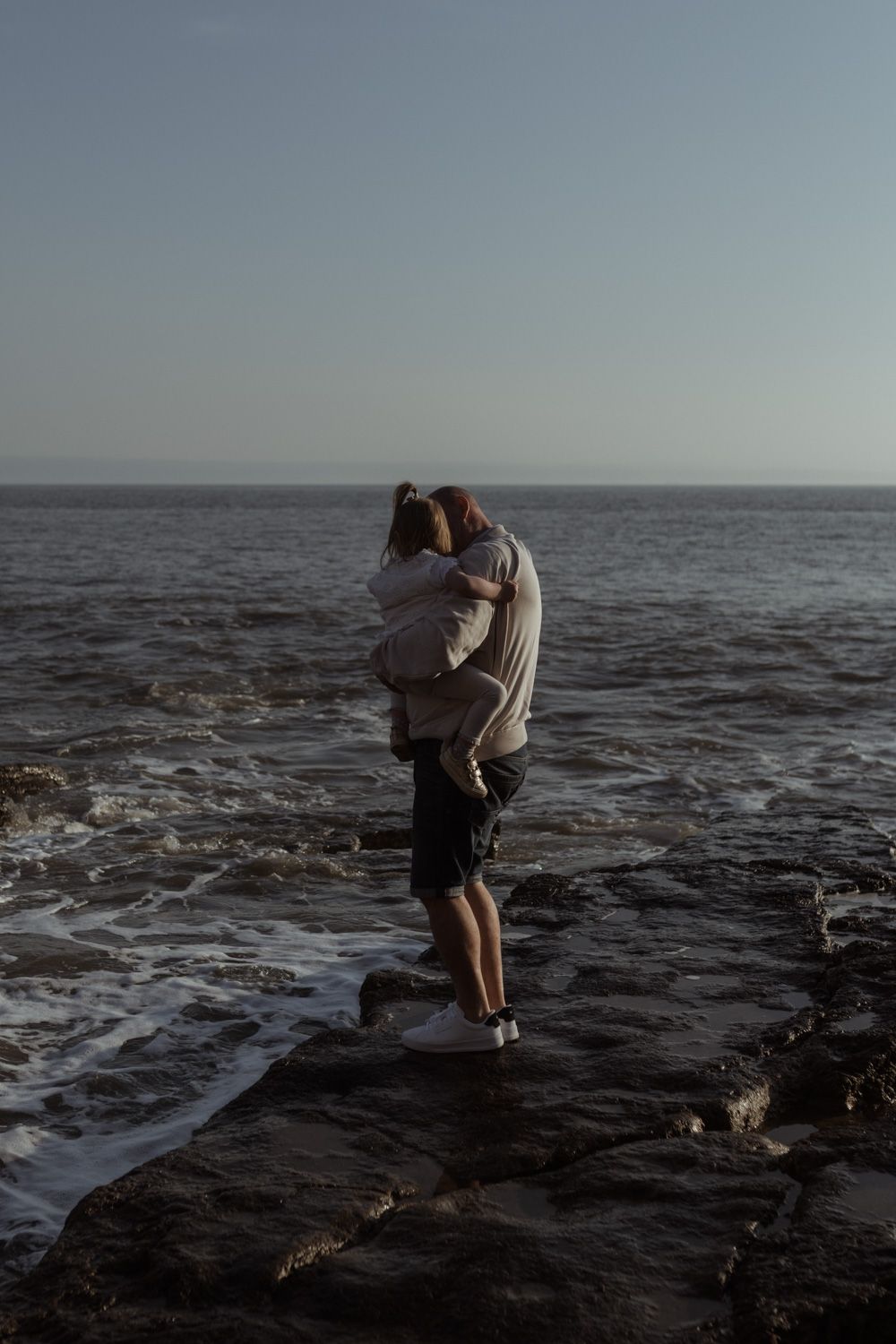 A silhouetted couple embraces on rocky ocean shoreline at dusk.