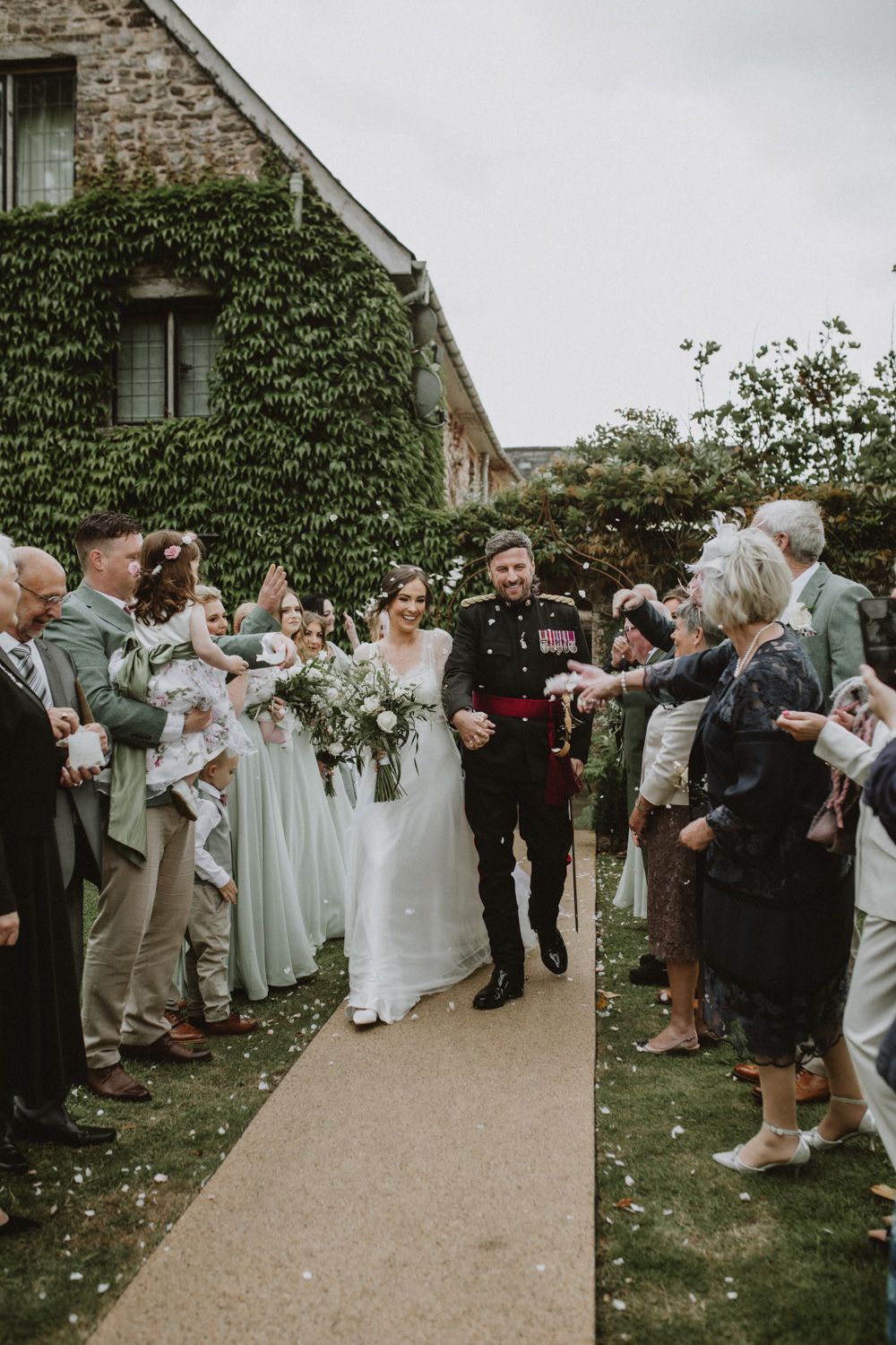 Wedding celebration in front of ivy-covered stone building as guests line pathway to celebrate the newlyweds.