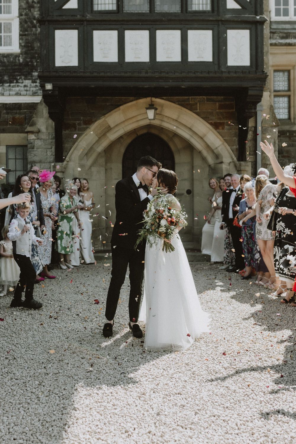 Wedding celebration exit under a stone archway with guests throwing confetti.
