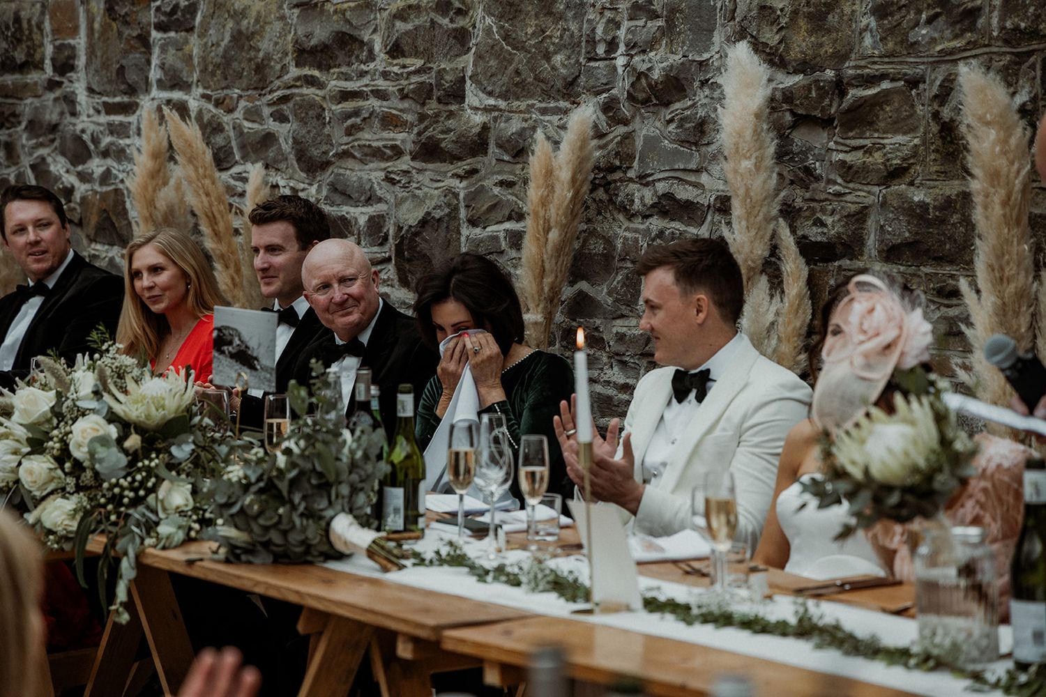 Wedding reception table with white floral centerpieces, pampas grass and guests gathered at a rustic stone wall venue.