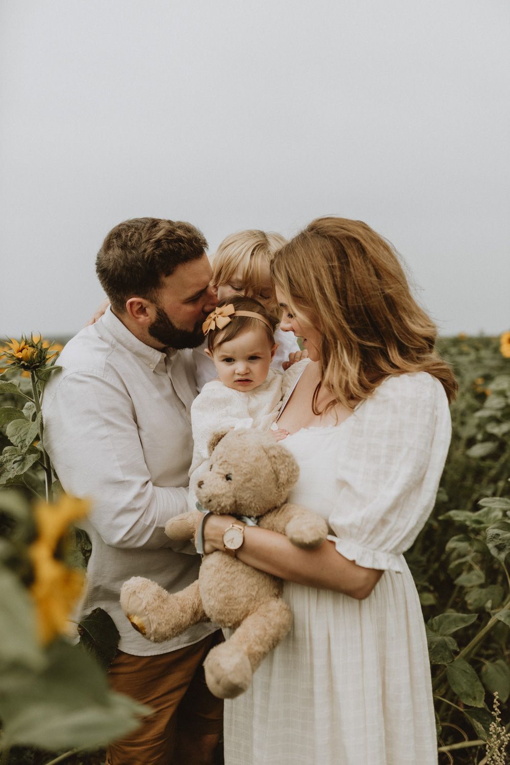 Sweet family moment in sunflower field with teddy bear and white clothing.