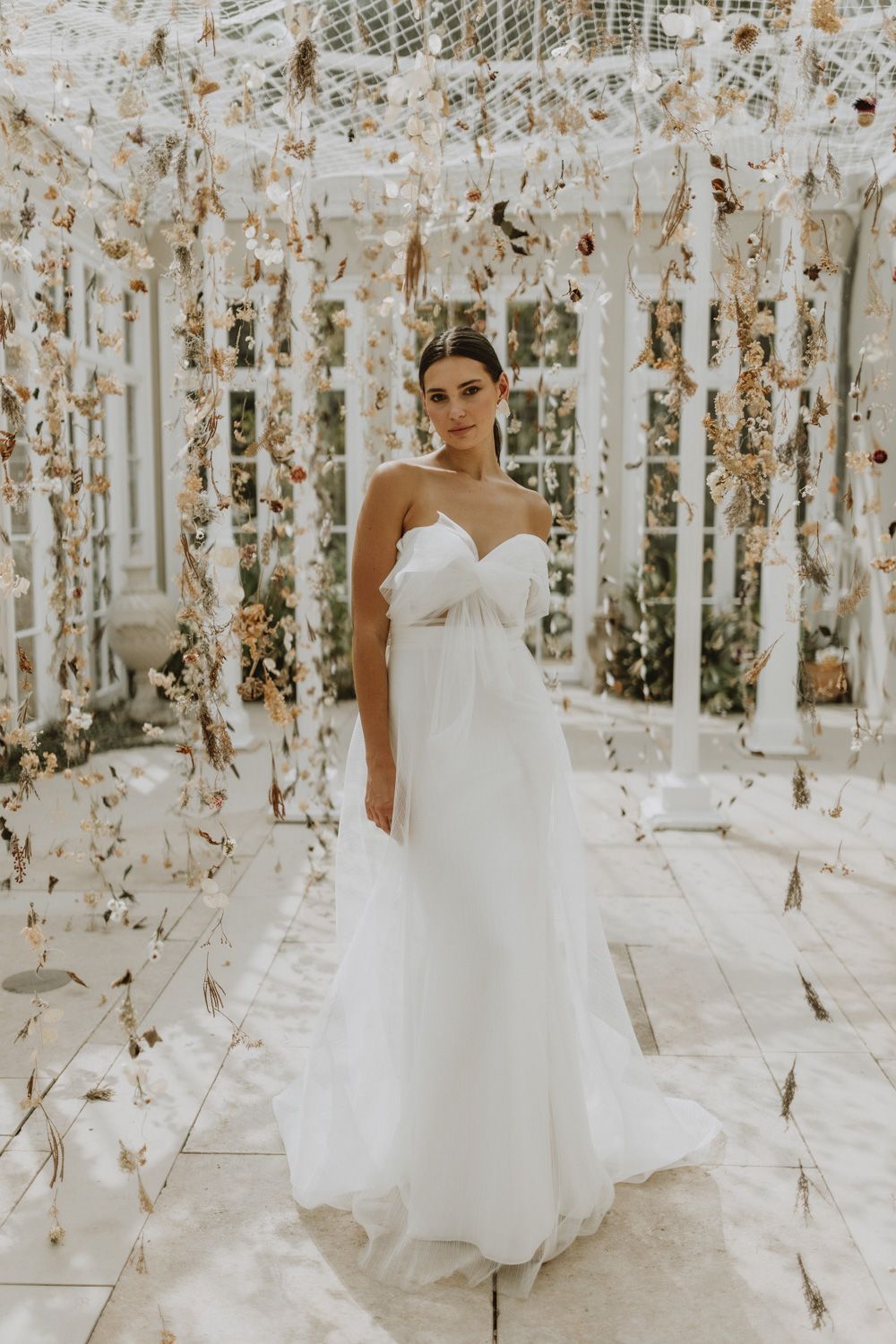 Ethereal bridal portrait in a white strapless gown within a floral-adorned conservatory setting.