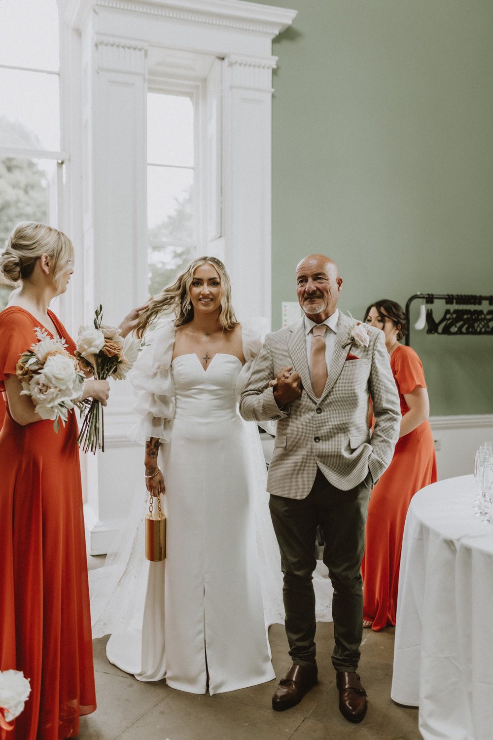 Wedding party in red dresses and light suits sharing emotional moment in bright room.