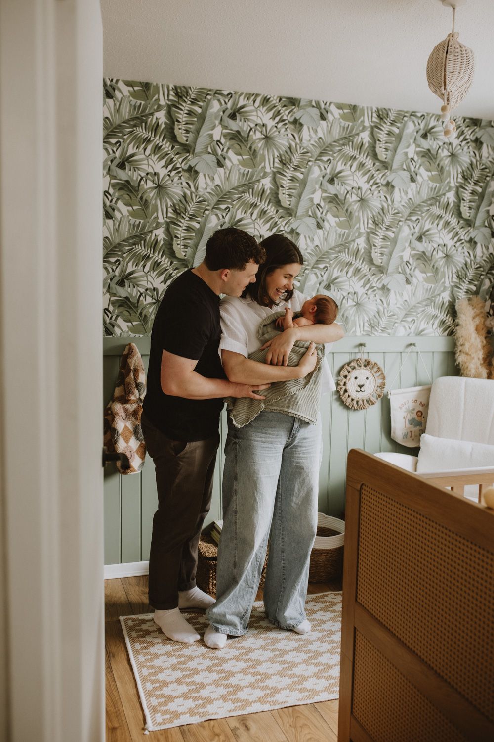 Modern nursery room with tropical wallpaper and family gathering around a changing table.
