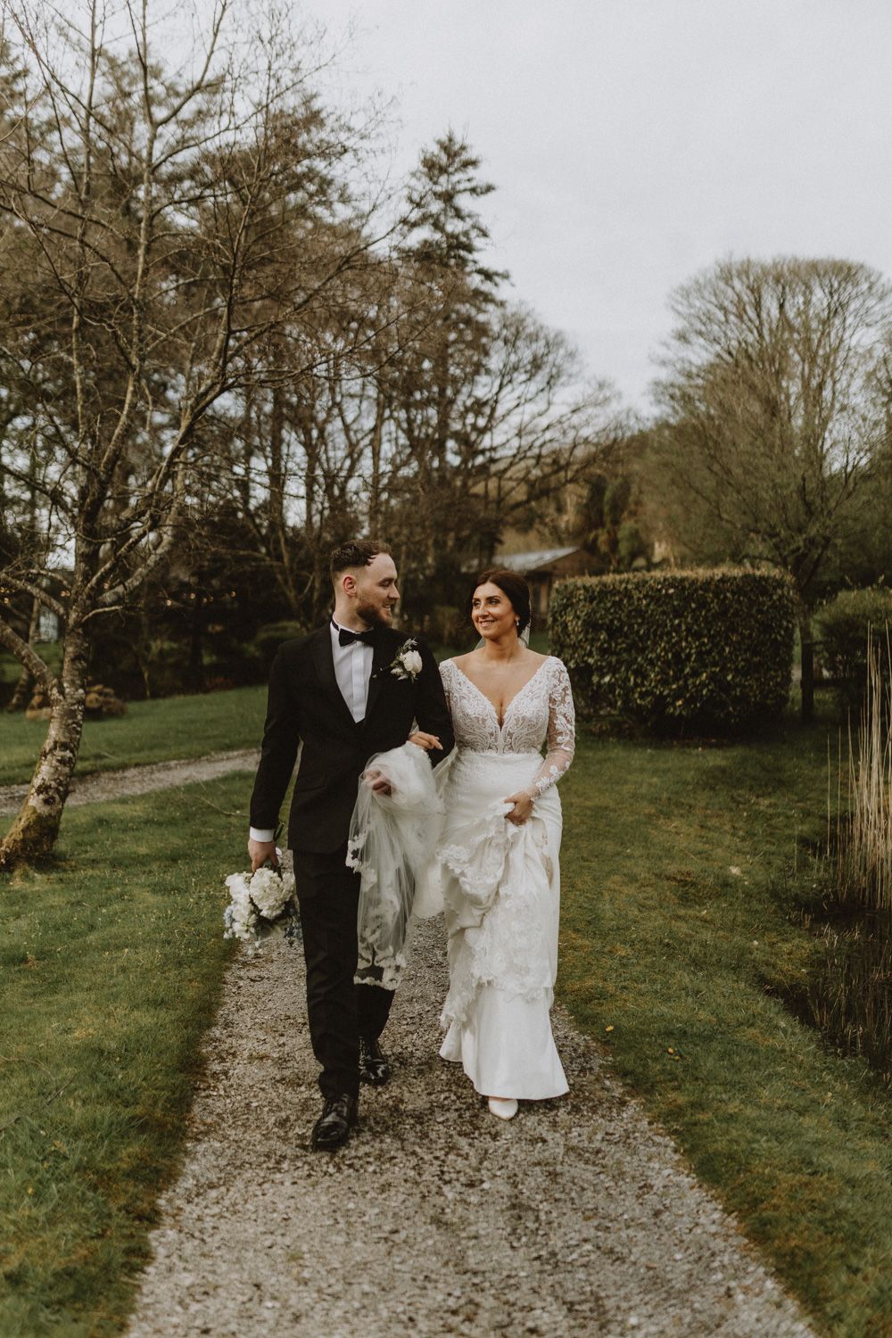 Newlyweds walking along a garden path surrounded by winter trees and greenery.