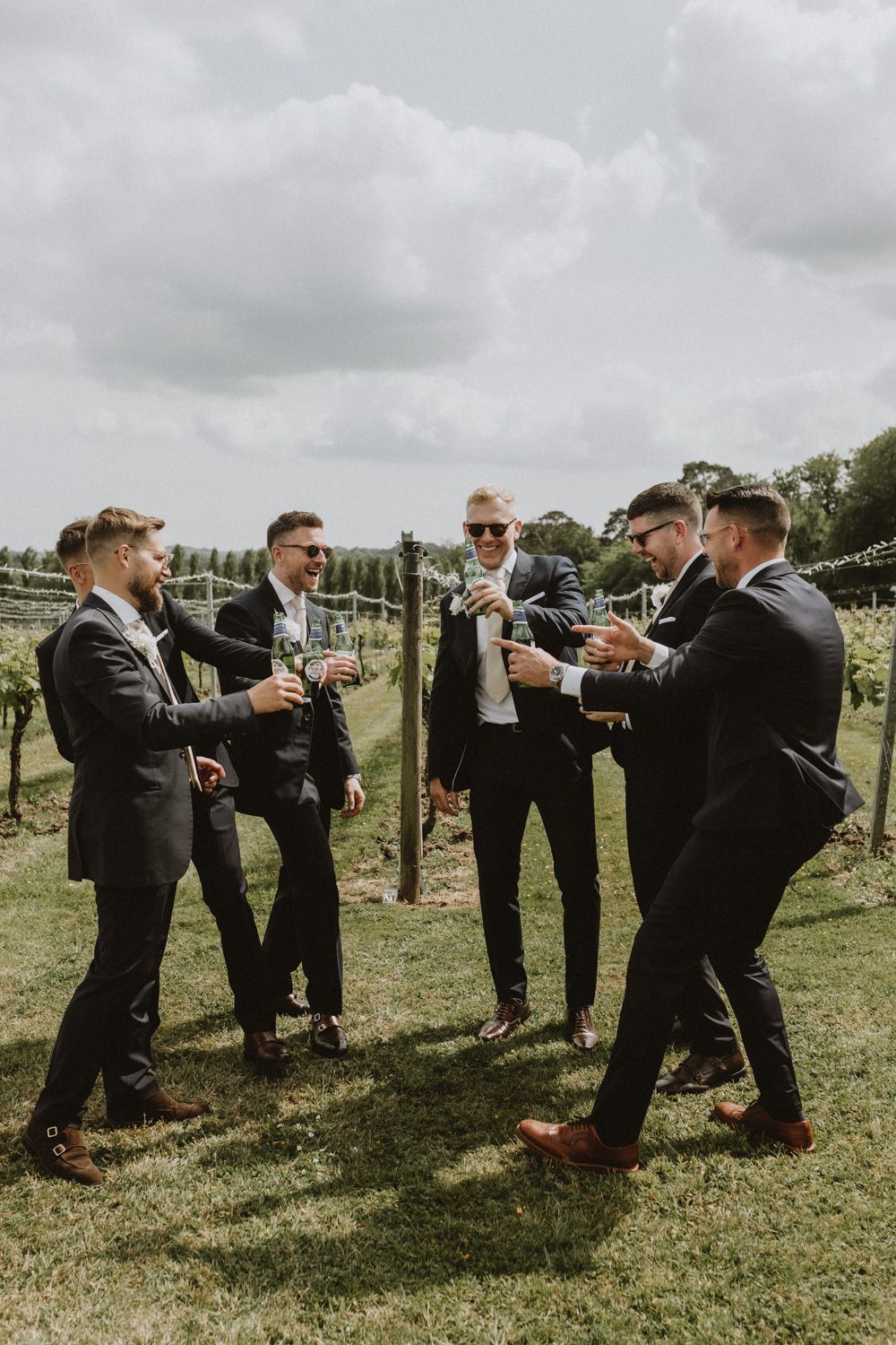 Groomsmen in black suits toast and celebrate together on vineyard grounds.
