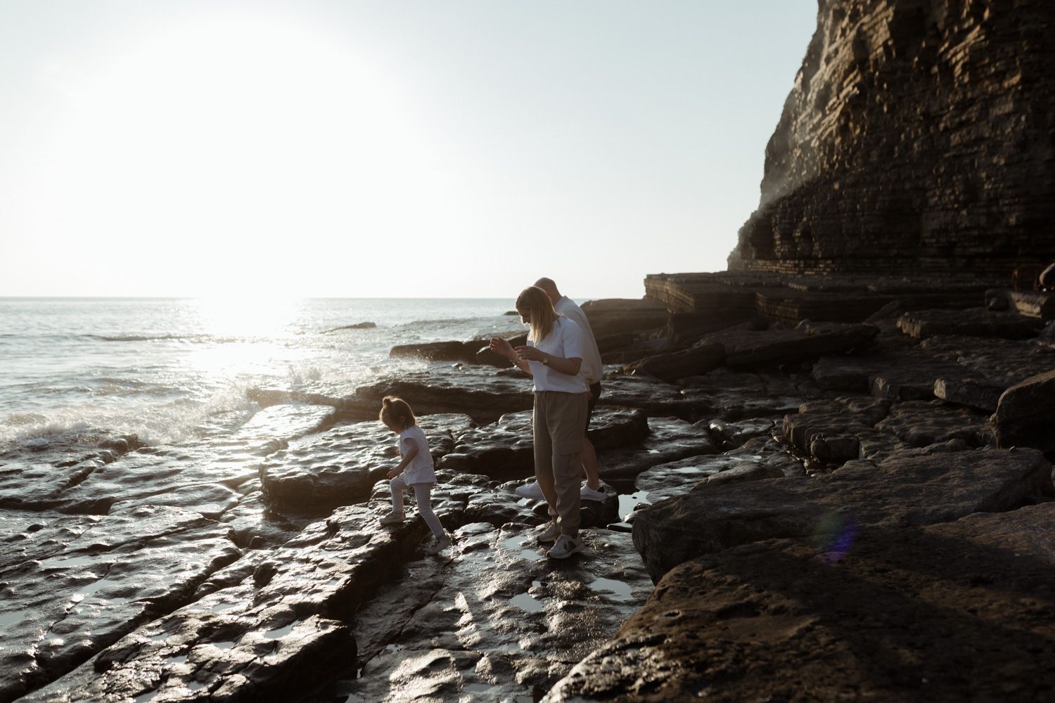 Figure stands on rocky coastal shoreline during golden hour with cliffs and ocean waves.