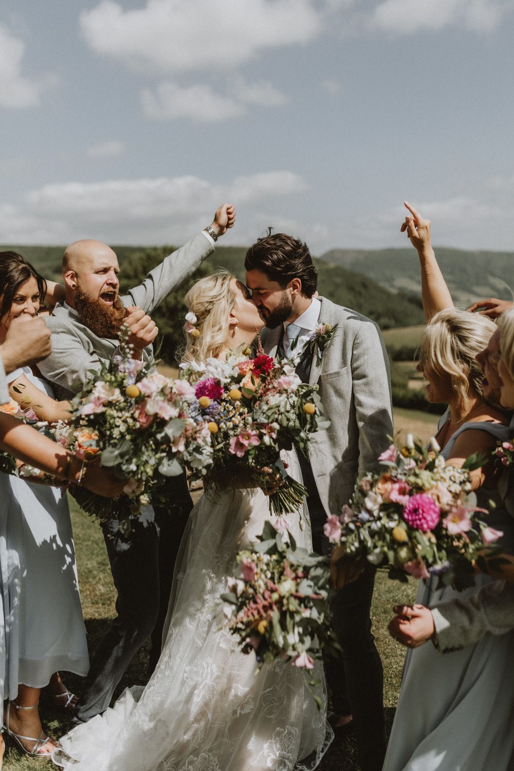 Wedding party celebrates joyfully outdoors with colorful bouquets and raised hands.