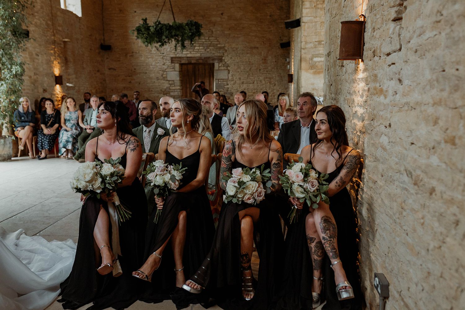 Bridesmaids in navy blue dresses holding white floral bouquets while standing against rustic stone wall at wedding ceremony.