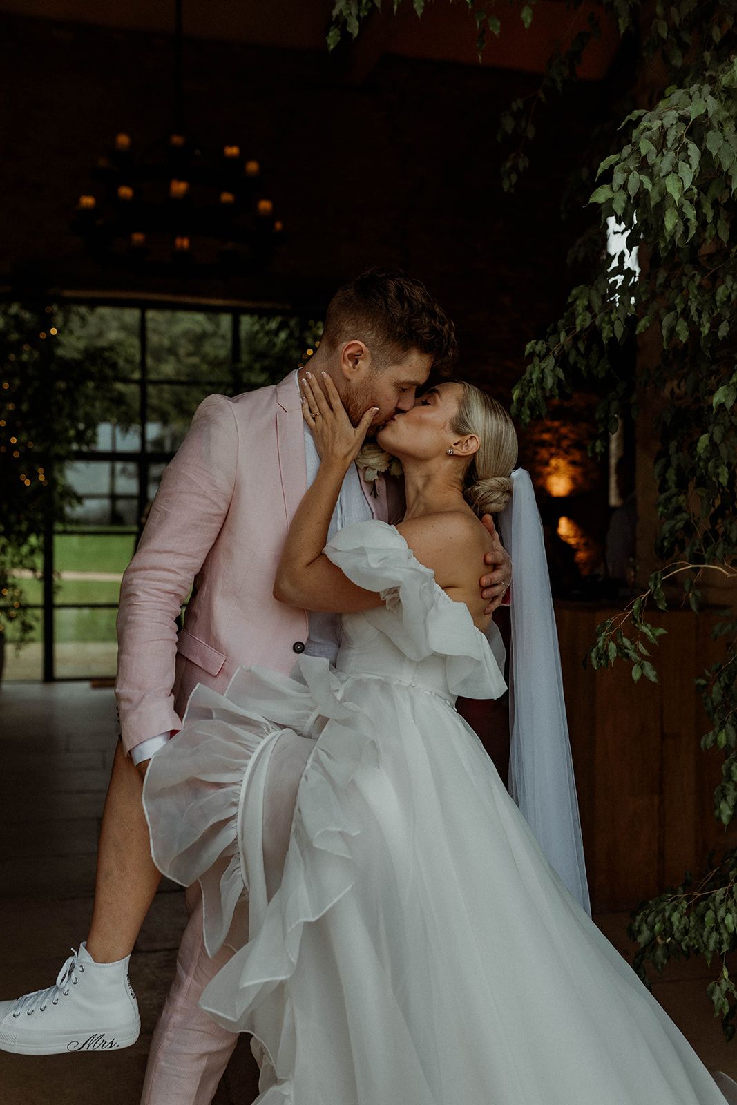 A romantic newlywed couple sharing an intimate kiss outdoors, with greenery and ambient lighting creating a dreamy atmosphere.