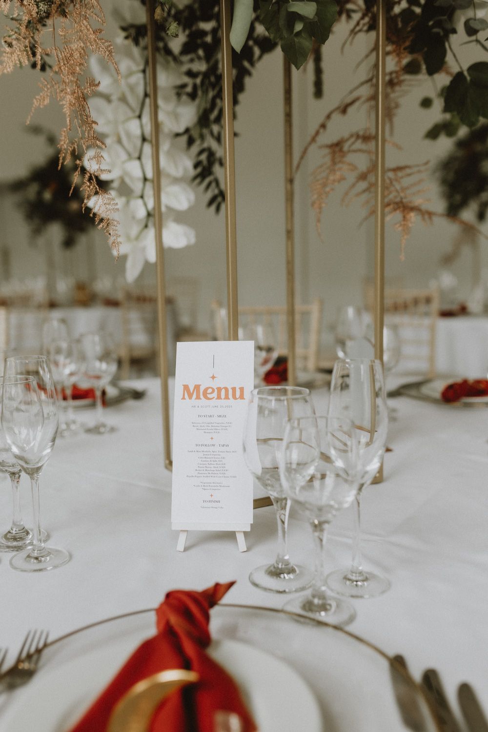 Elegant wedding table setting with menu card, wine glasses and red napkin accents.