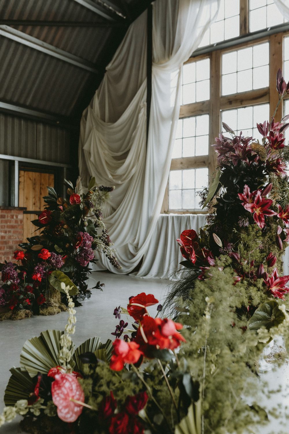 Dramatic white fabric draping and red floral arrangements decorate an industrial-style venue space.