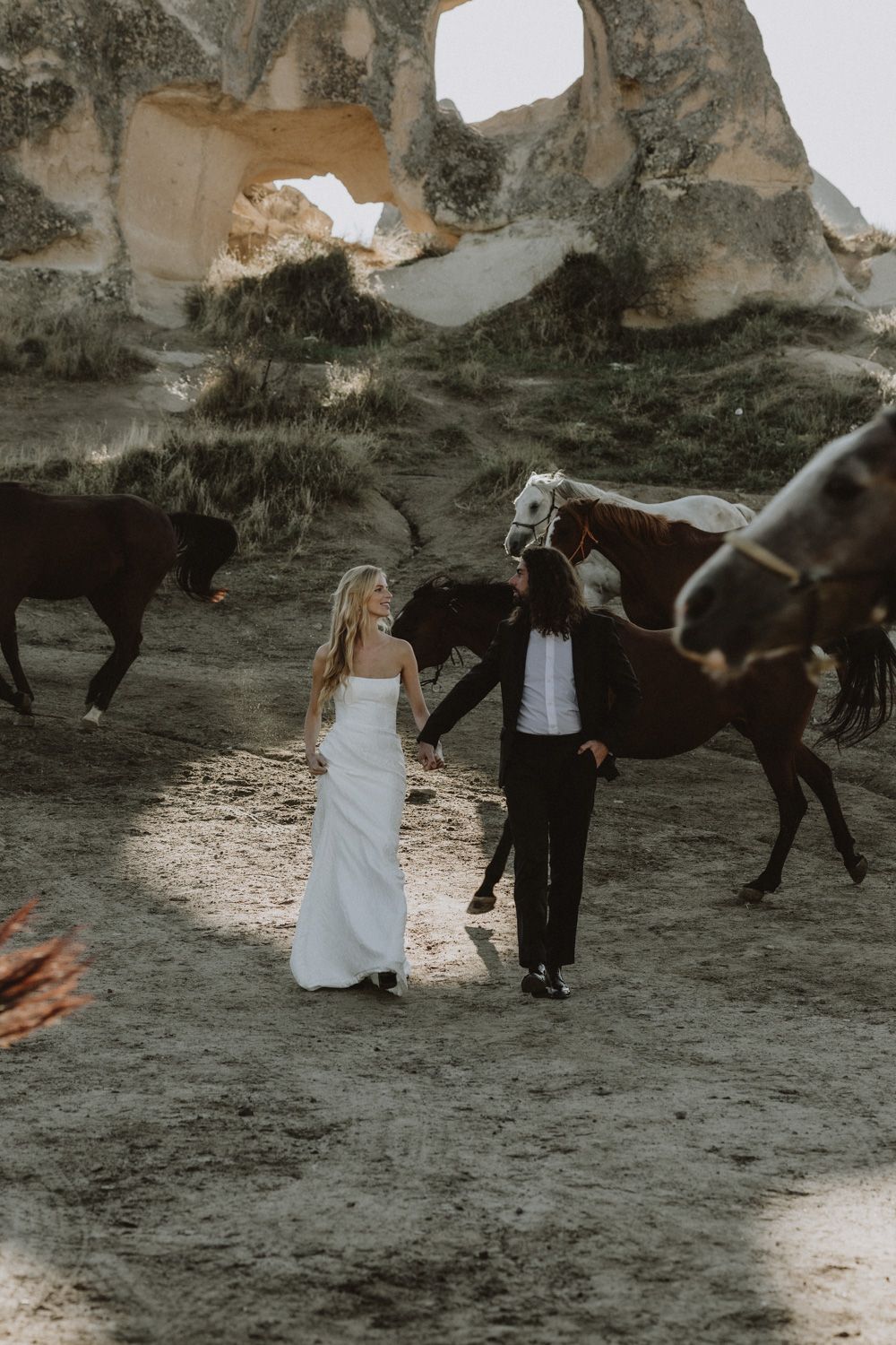 A wedding couple poses for photos with horses against a dramatic rocky backdrop.