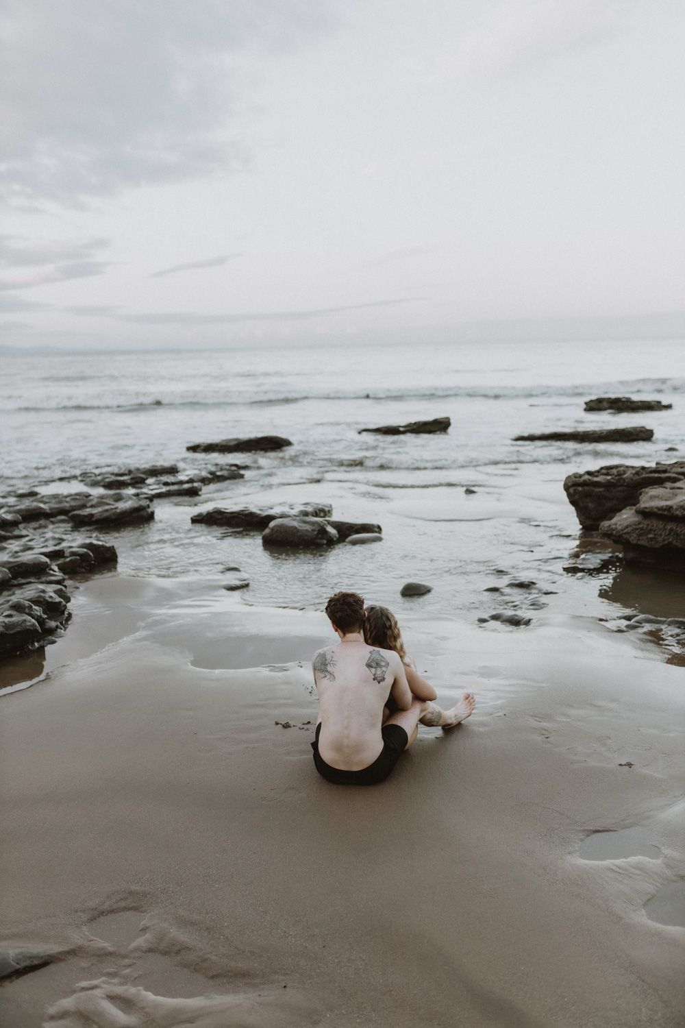 Couple sits together on a rocky beach at sunset with waves crashing against the shore.