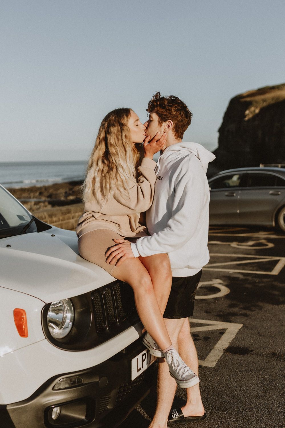 Romantic couple sharing kiss while leaning against white car at coastal viewpoint.