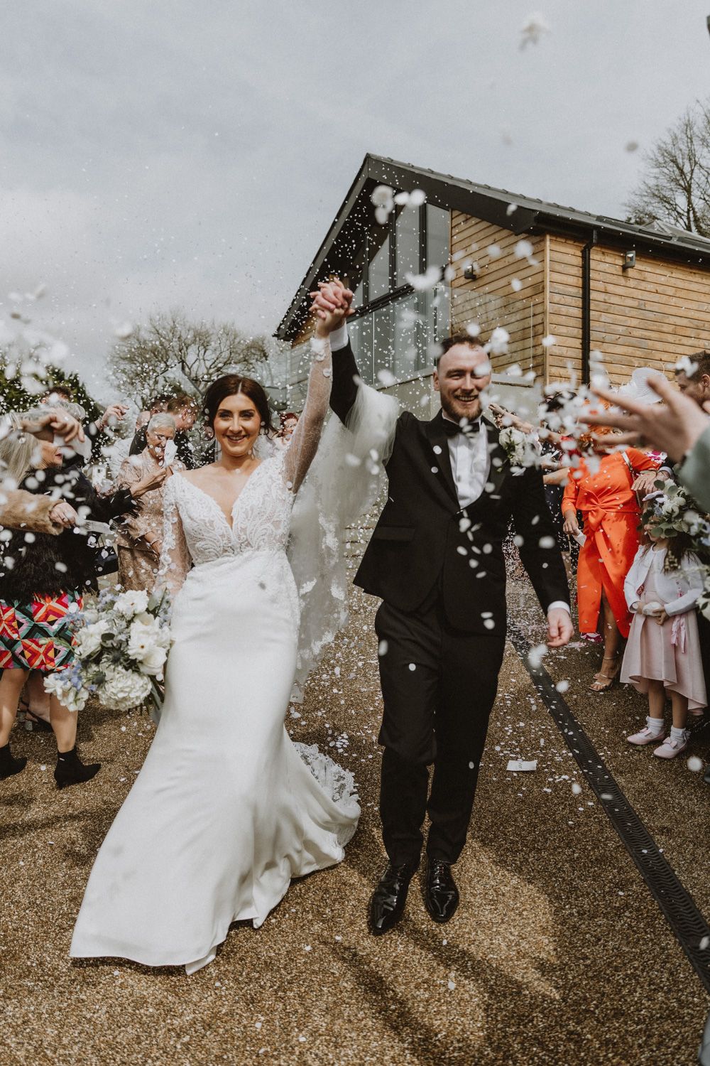 Wedding guests celebrate by throwing confetti at a newlywed couple outside a rustic venue.