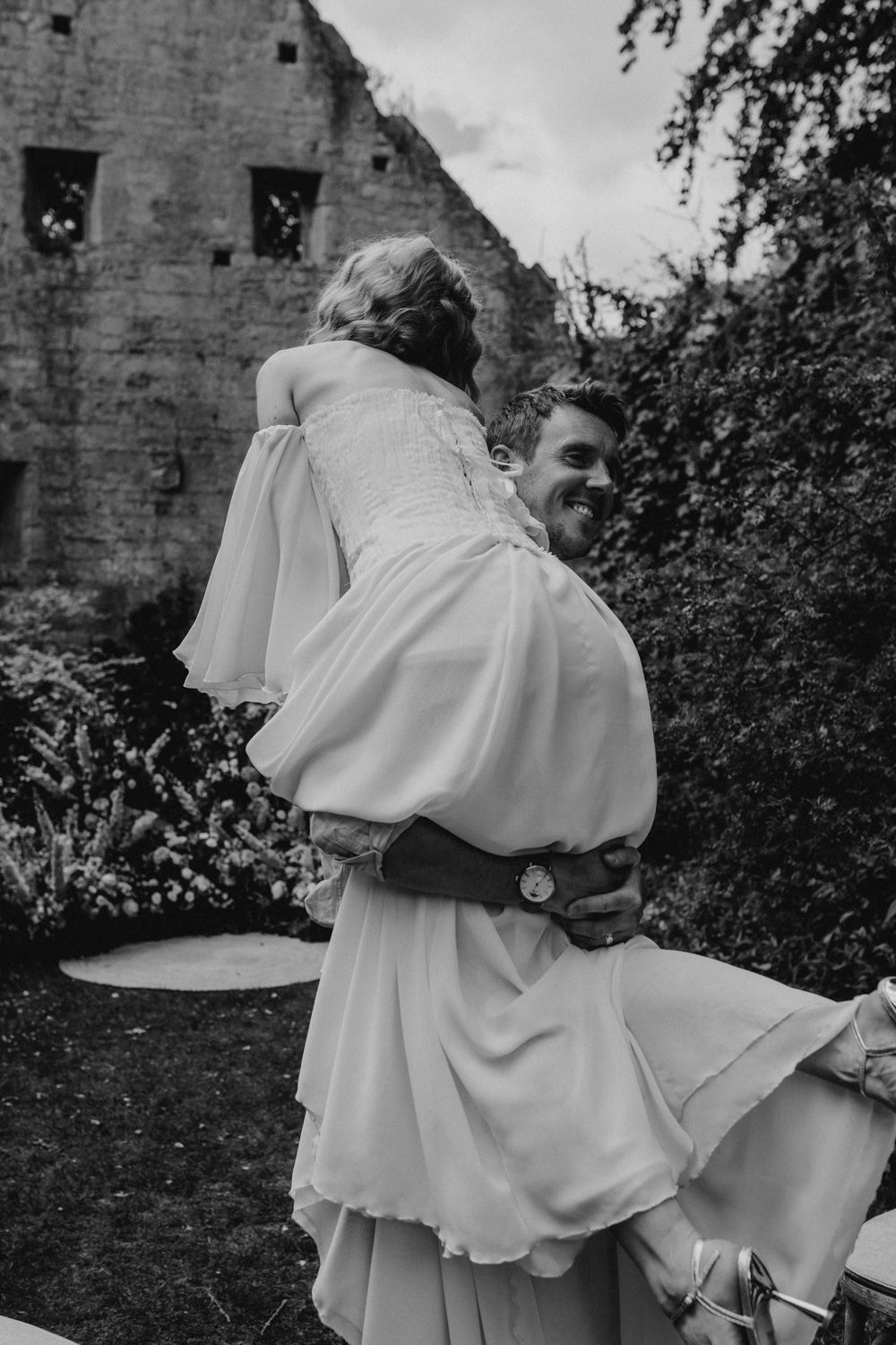 Black and white wedding photo of playful couple sharing a joyful moment in front of stone ruins.