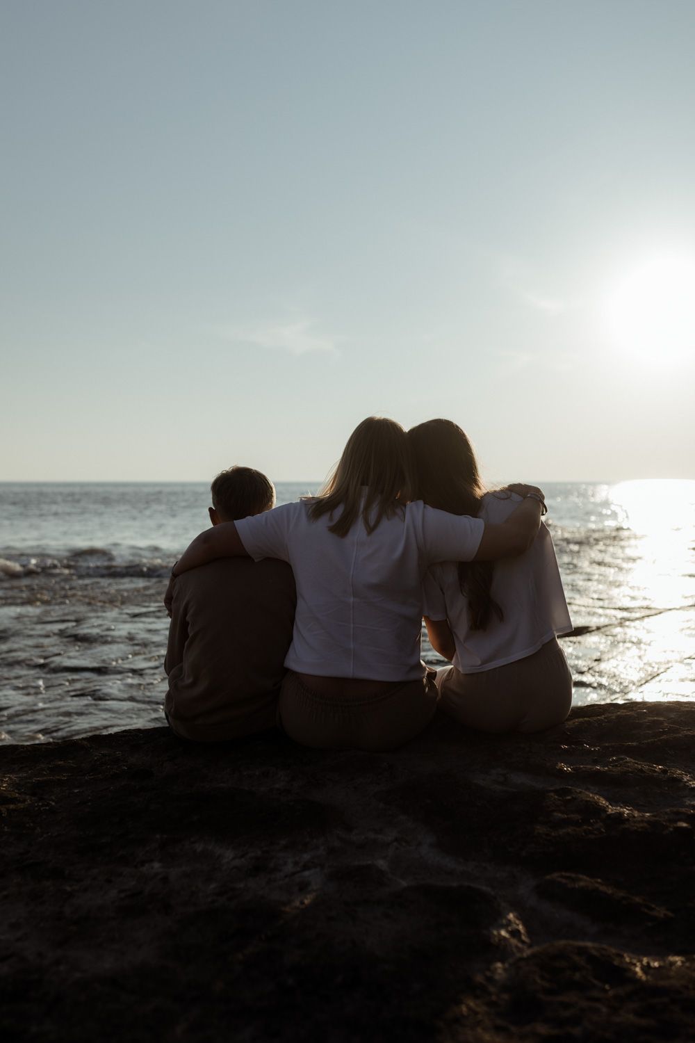 Three silhouettes sit together watching sunset over ocean from rocky cliff.