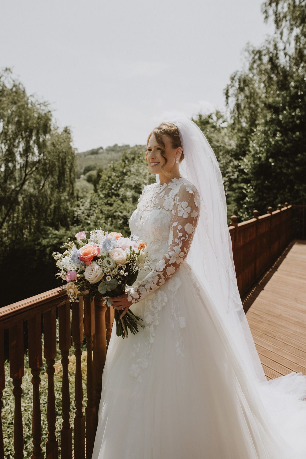 Beautiful bride in lace gown with flowing veil standing on wooden deck.