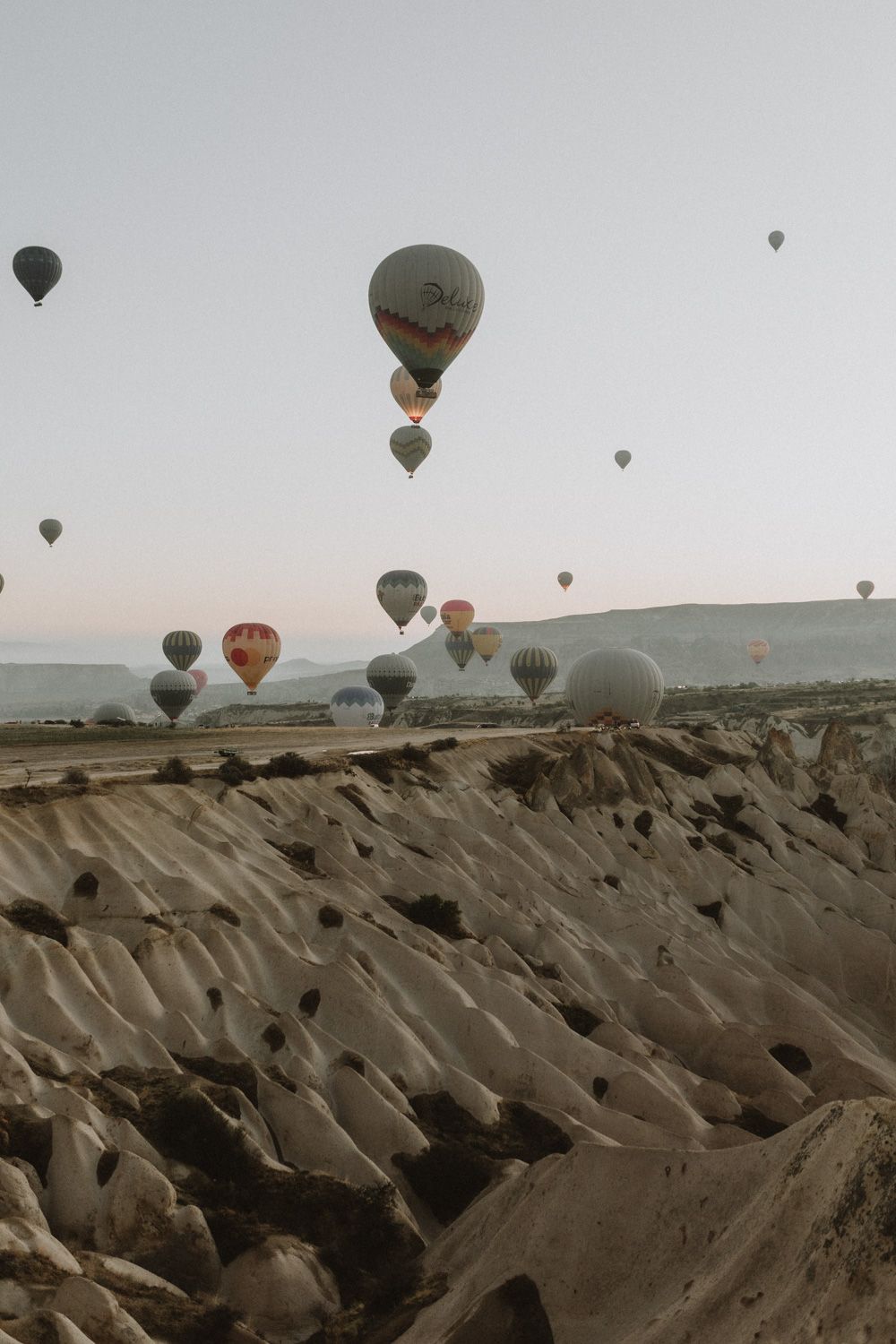 Panoramic view of hot air balloons floating over rocky Cappadocia landscape during sunrise.