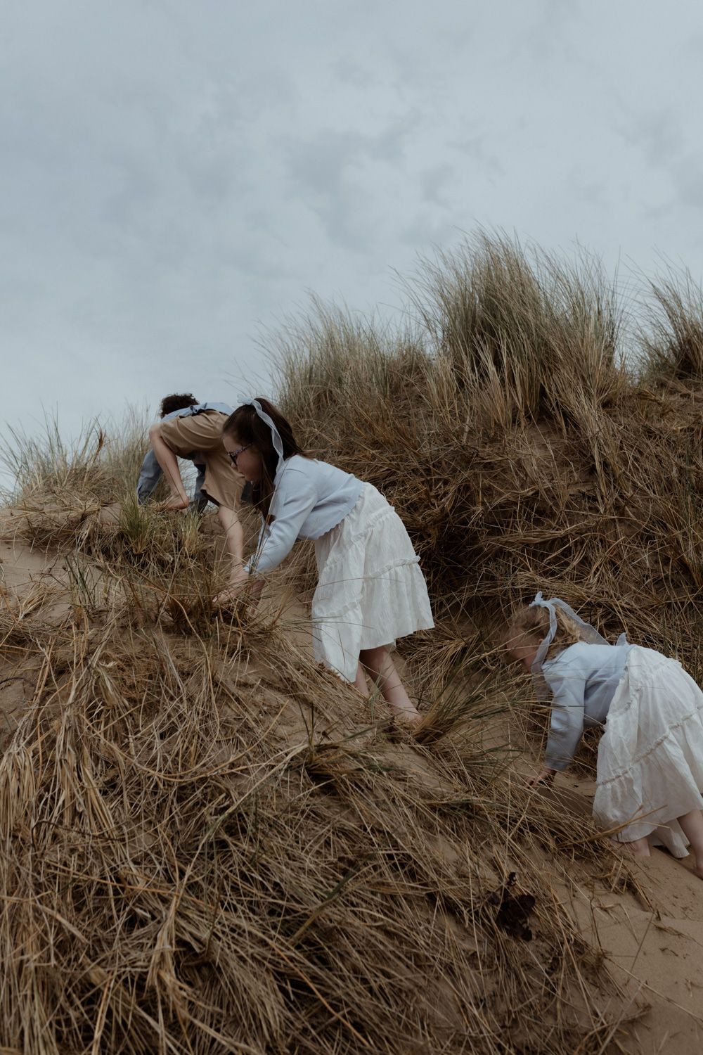 Two people in white dresses climb sand dunes covered in beach grass.