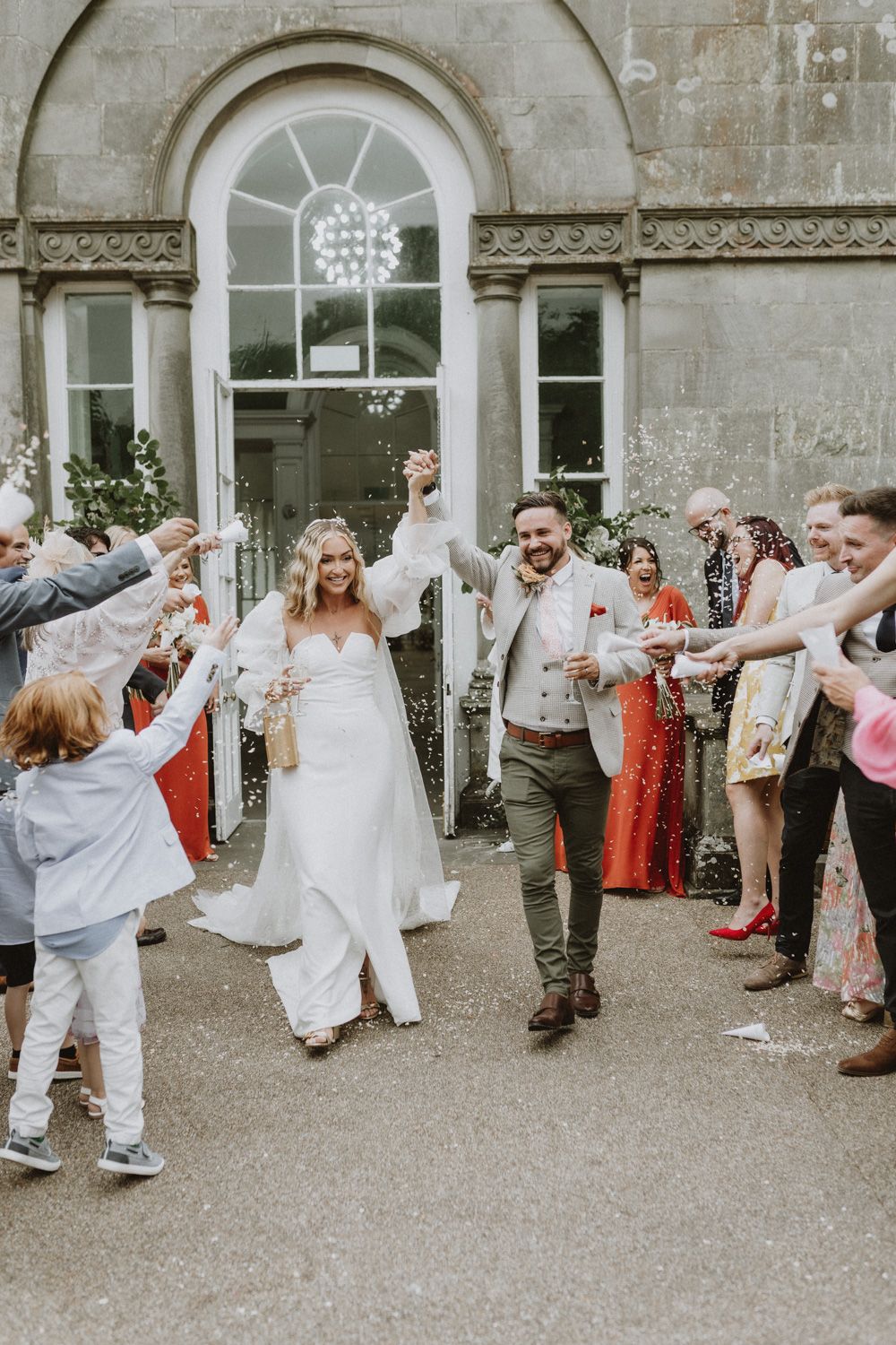Wedding celebration outside stone building as guests shower couple with confetti.