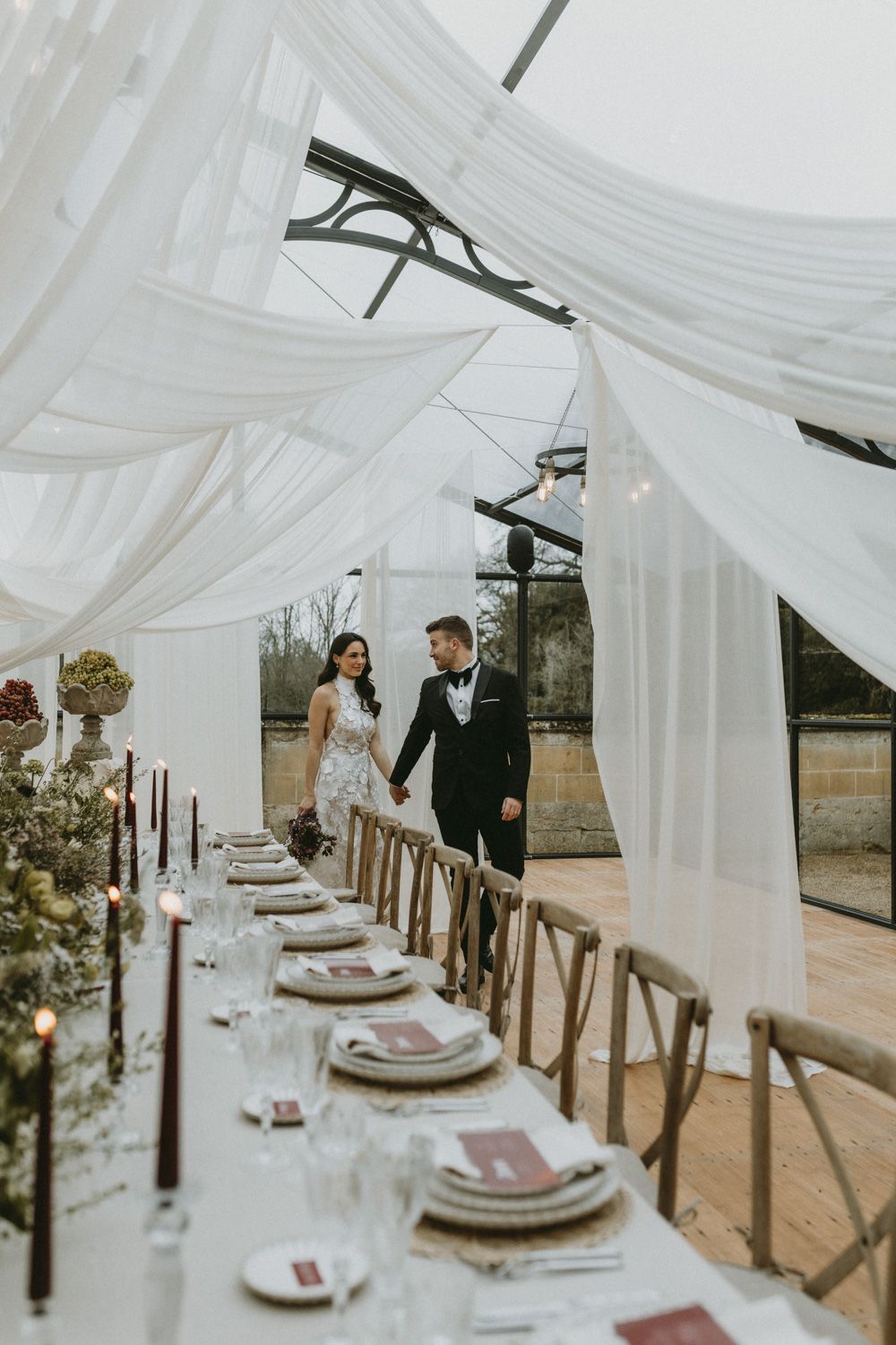 Long wedding reception table with white draping overhead, wooden chairs, and elegant place settings.