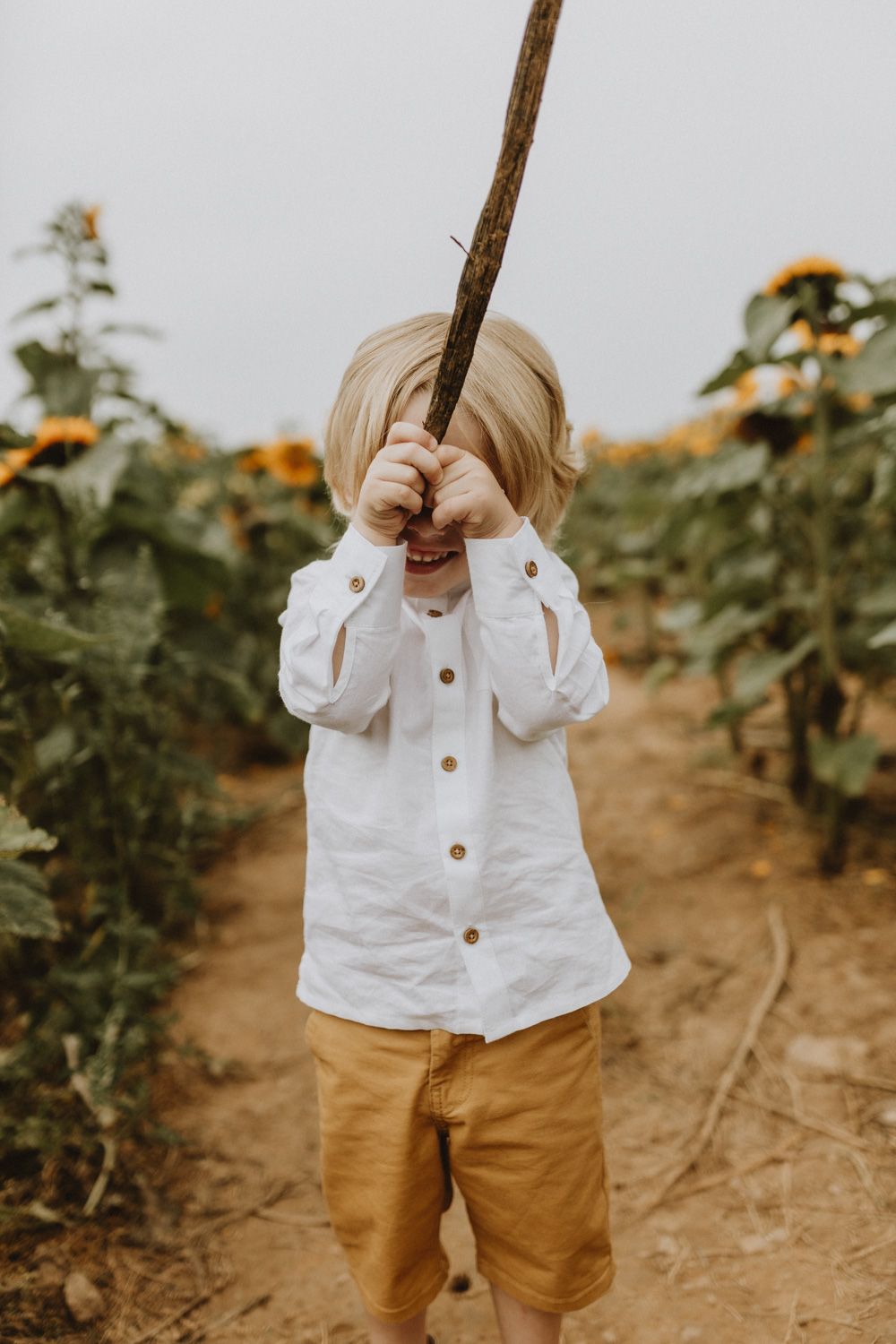 Child in white shirt and khaki shorts playing with stick in sunflower field.