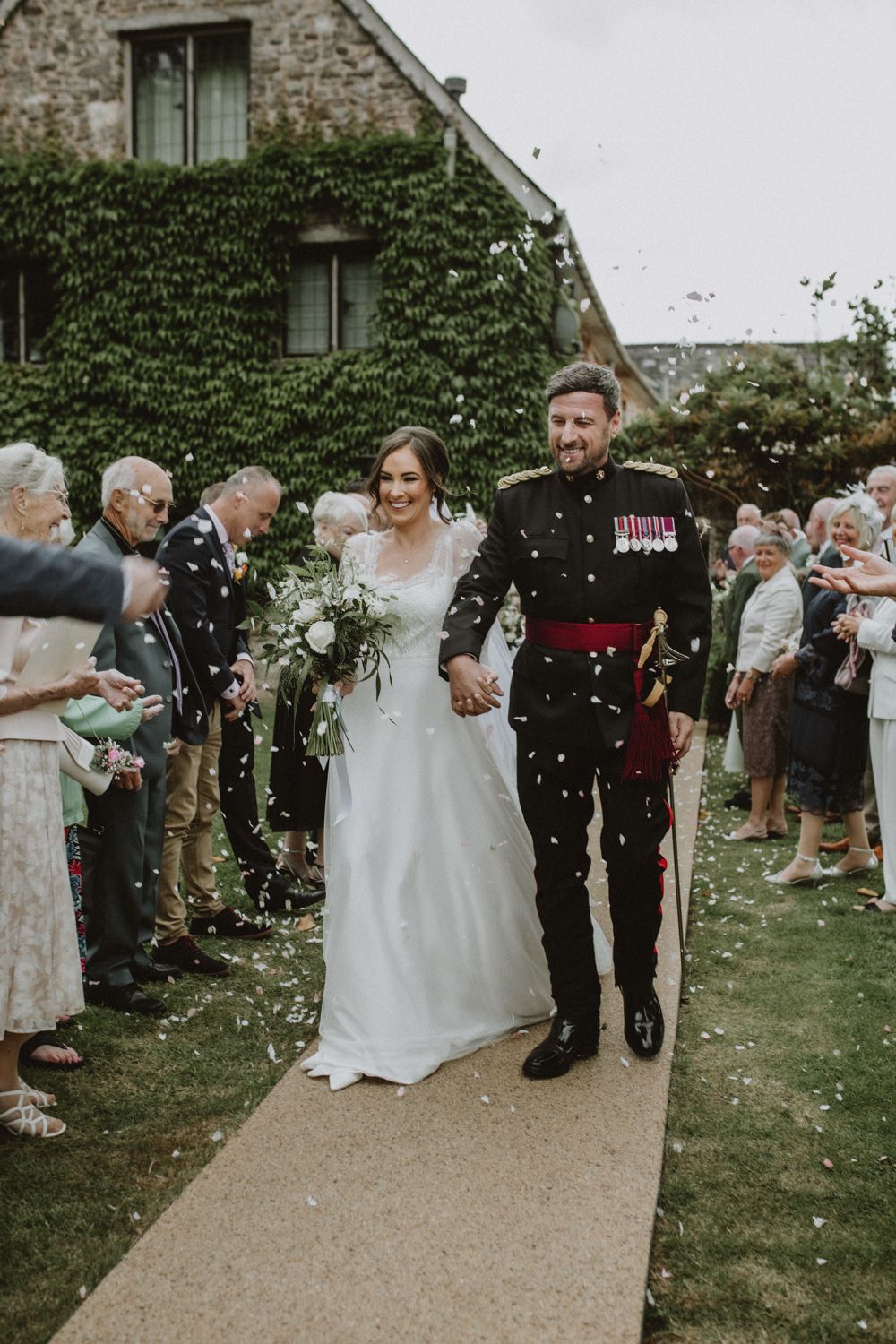 A wedding celebration outside a stone house covered in ivy as guests throw confetti on the pathway.