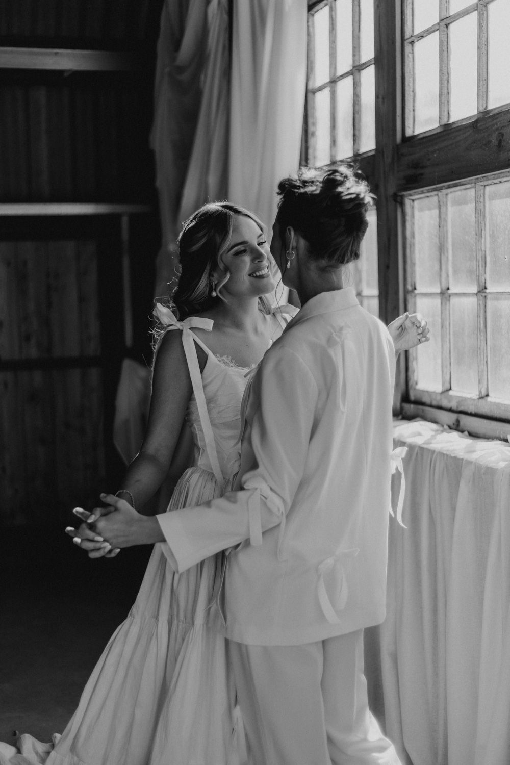 A black and white portrait of a tender wedding moment by a sunlit window.