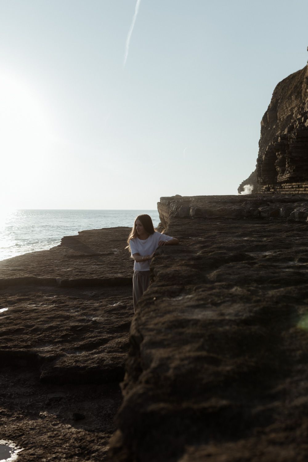 Person stands on dramatic rocky cliff edge overlooking ocean at sunset.