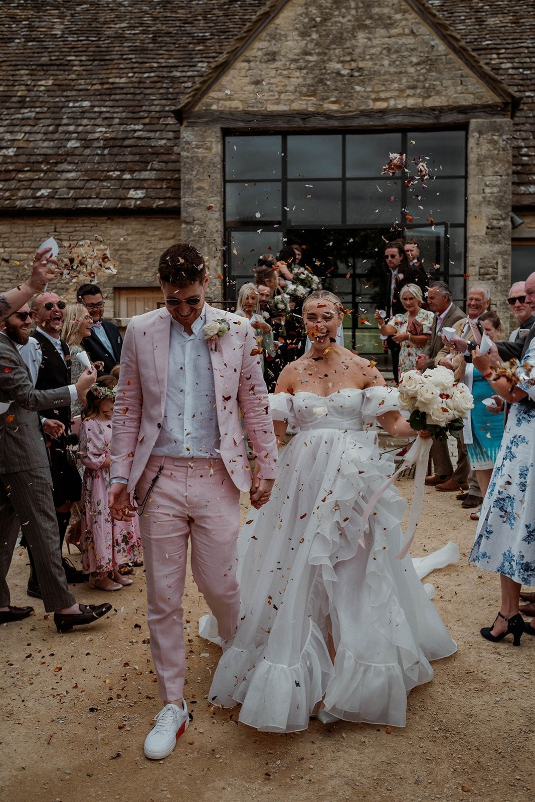 Wedding celebration with guests showering confetti on couple in pink suit and white gown outside stone building.