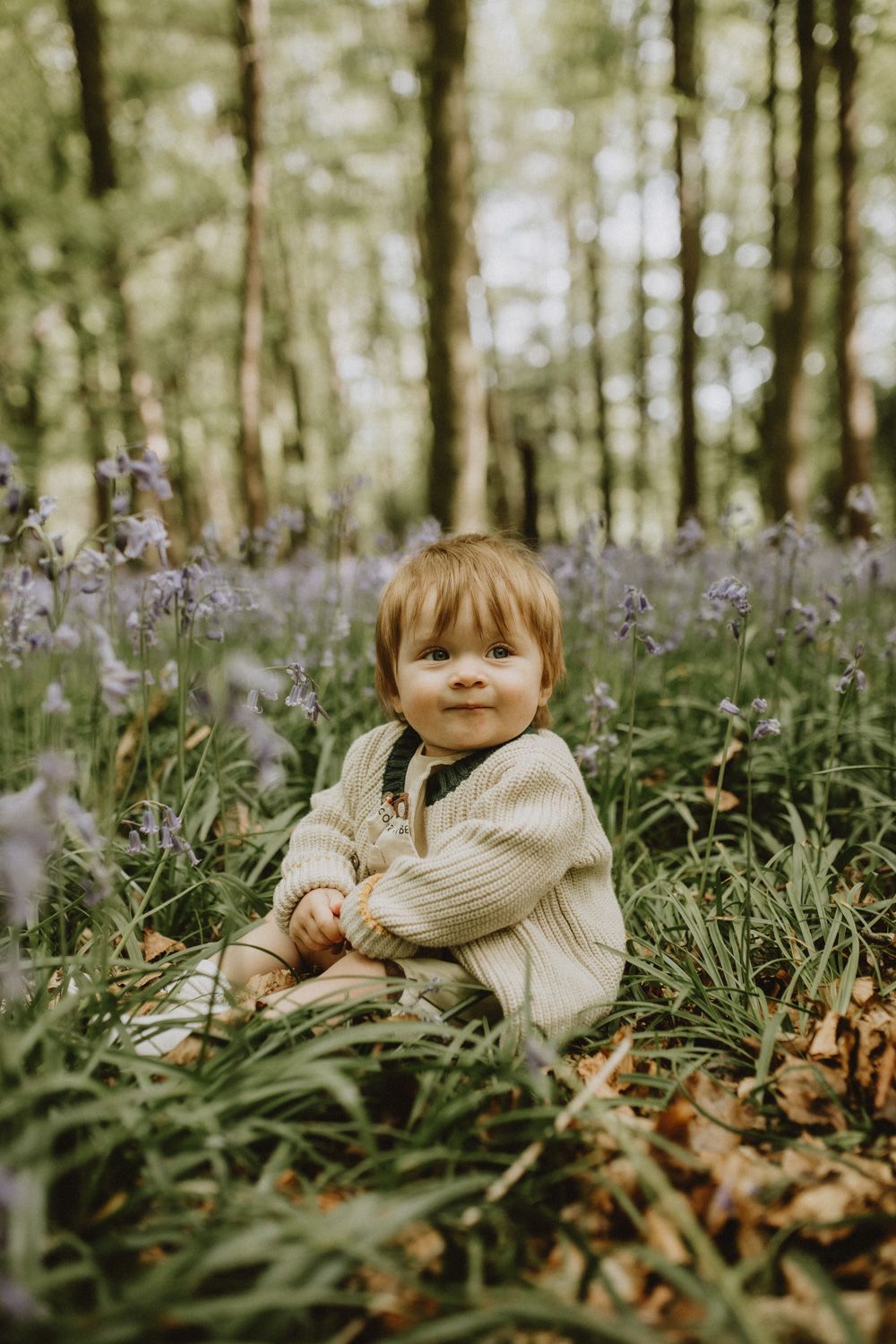 Child lying in field of purple wildflowers wearing beige sweater among woodland trees.
