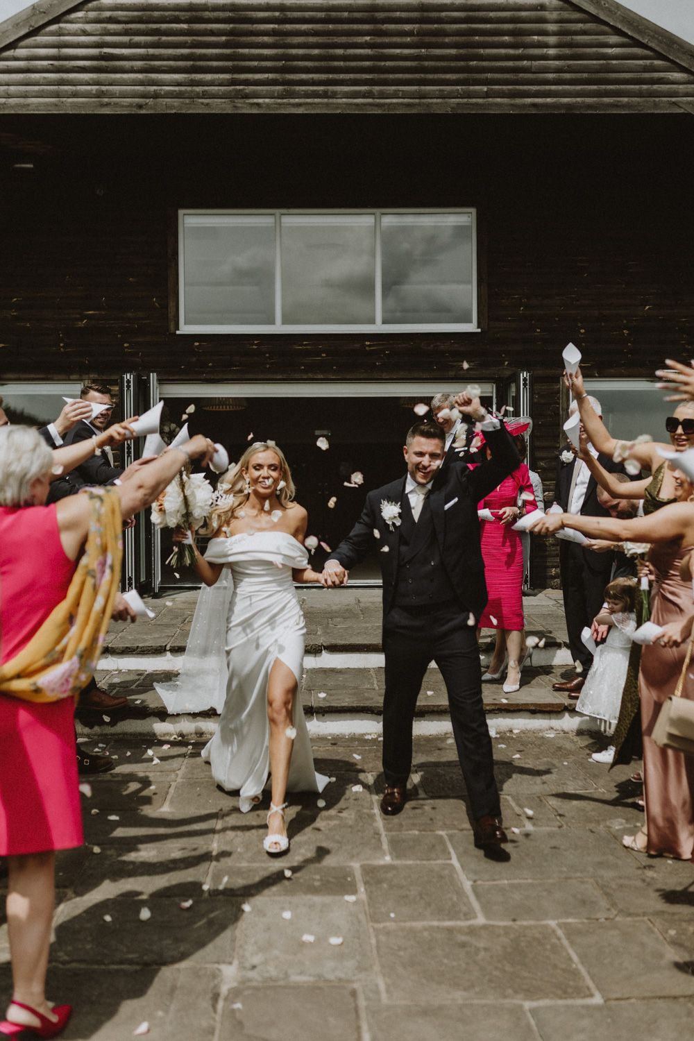 Joyful wedding exit with guests throwing confetti outside rustic venue building.