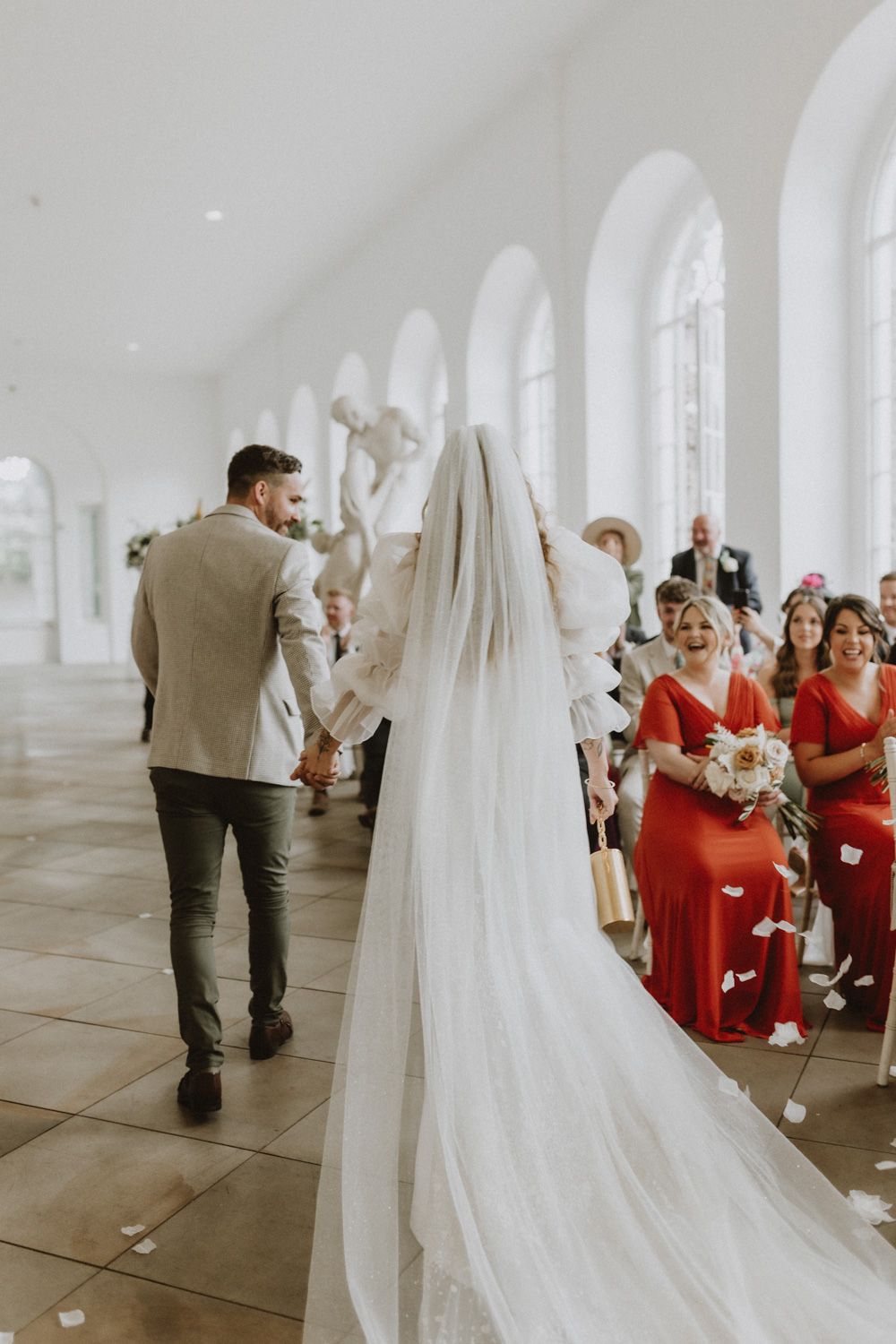 Wedding ceremony in elegant white chapel with guests in red dresses watching emotional moment.
