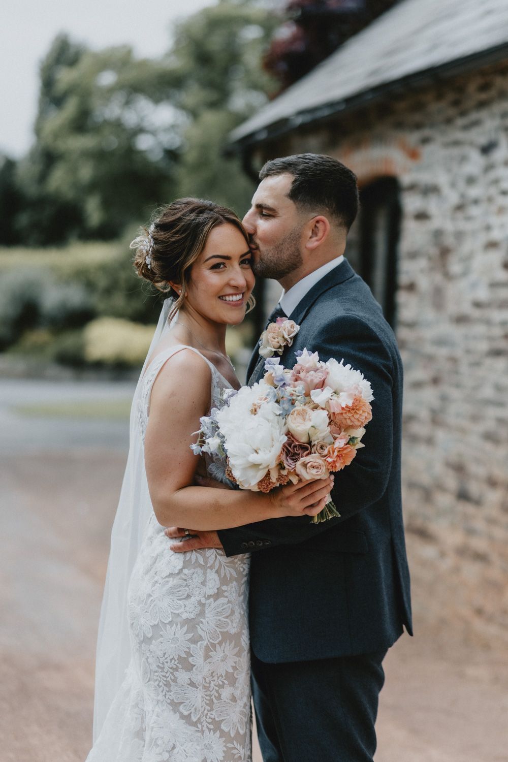 A romantic wedding portrait showing a couple embracing with a beautiful peach and white floral bouquet against a rustic stone wall.