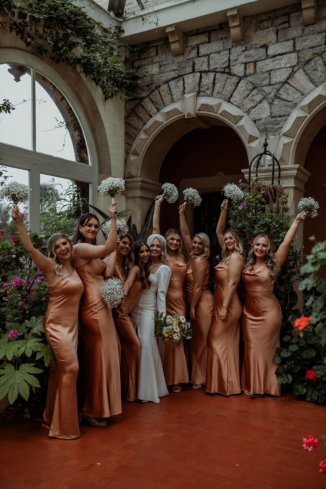 A wedding party in matching copper-colored dresses poses together in an elegant stone-walled courtyard with climbing vines.