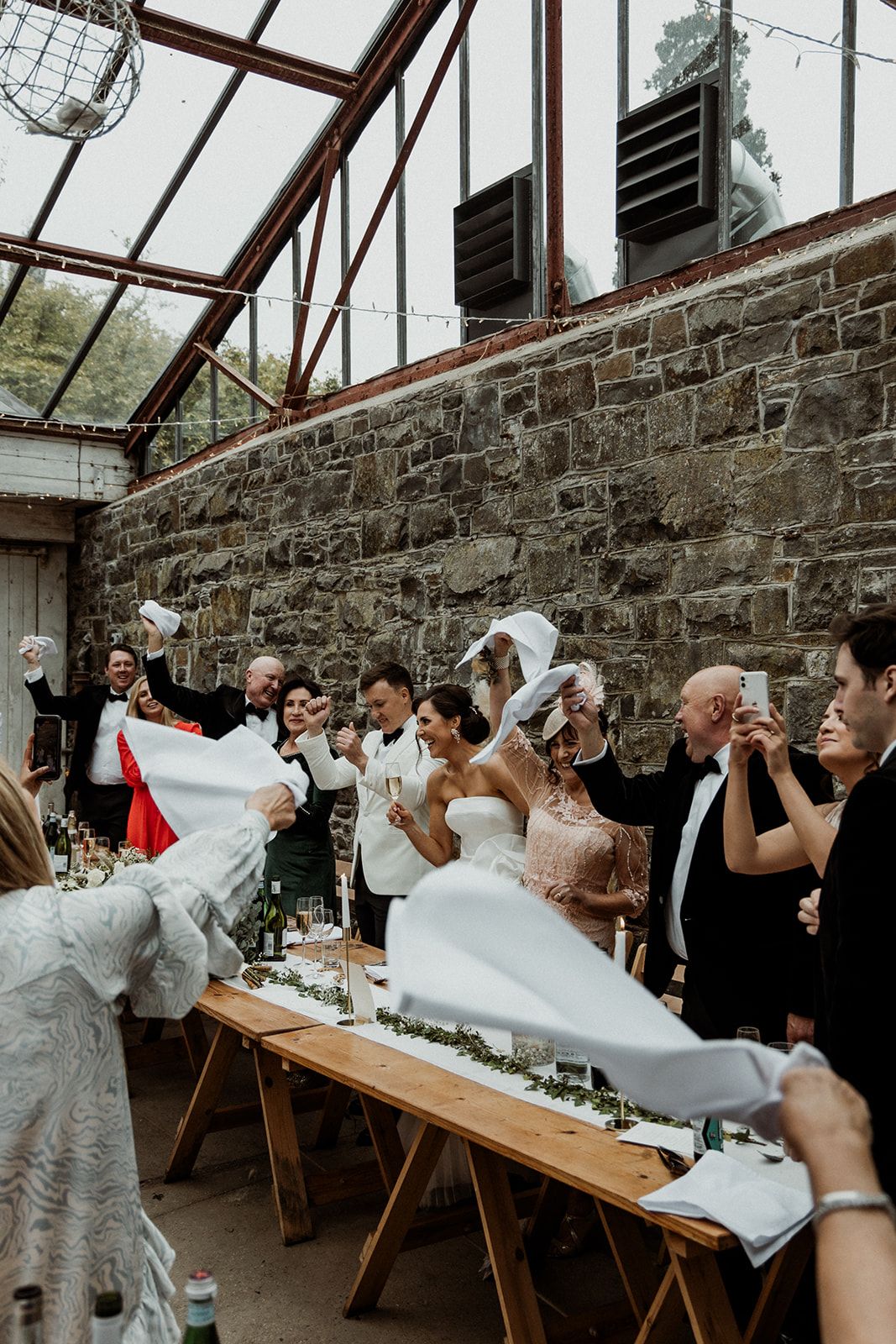 Wedding guests participate in a synchronized drinking game at long wooden tables against a rustic stone wall.
