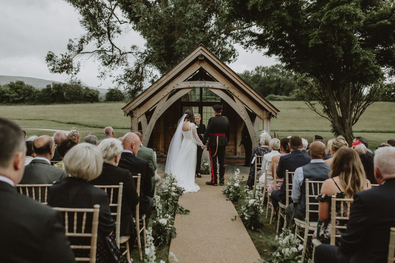 Rustic wooden outdoor wedding ceremony arch decorated with greenery on a scenic golf course with seated guests.