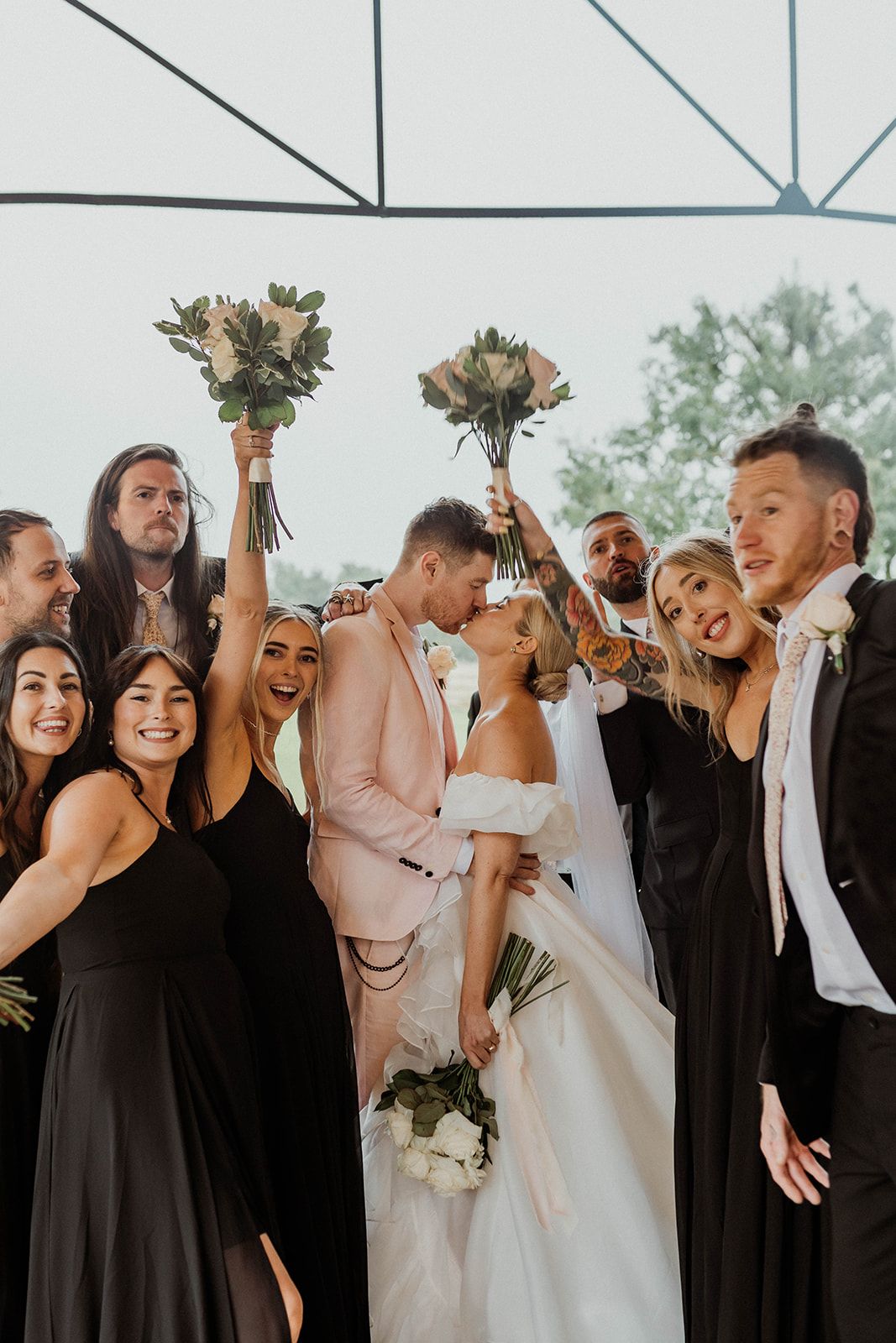 A joyful wedding party celebrates under a white tent, with bridesmaids in black dresses holding bouquets.