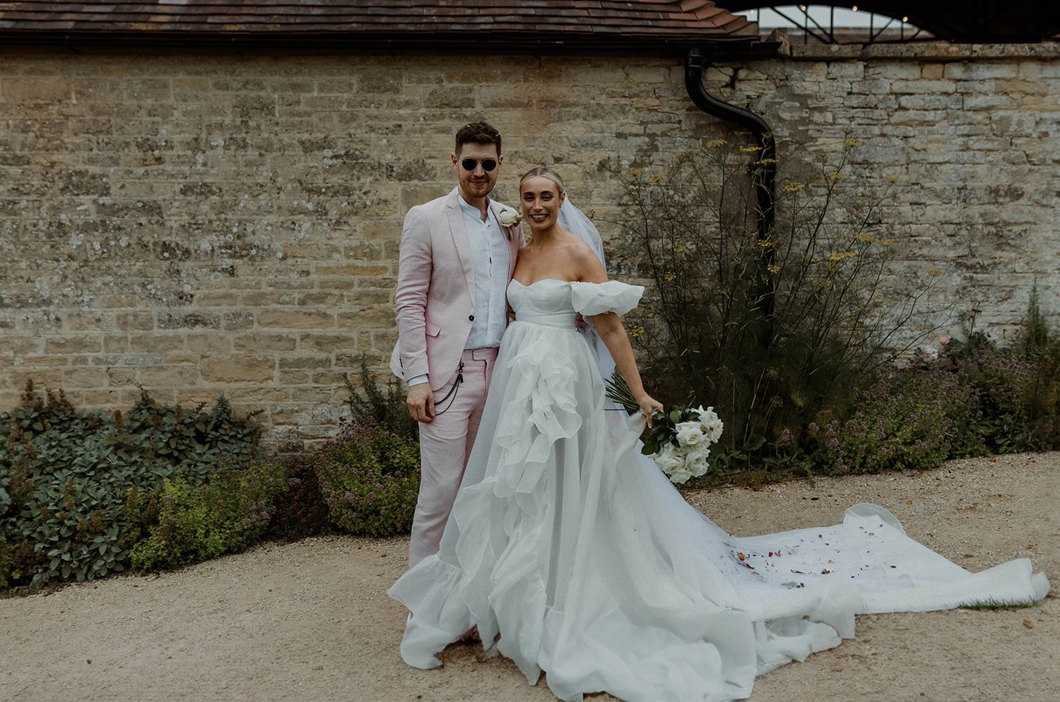 Elegant wedding couple poses together in front of rustic brick wall with flowing white dress and pink suit details.