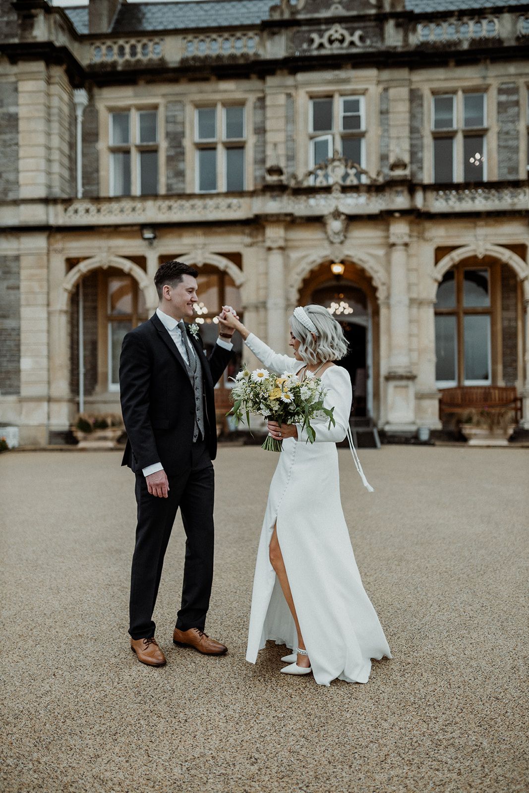 An elegant wedding couple poses in front of a grand historic stone building with ornate architecture.