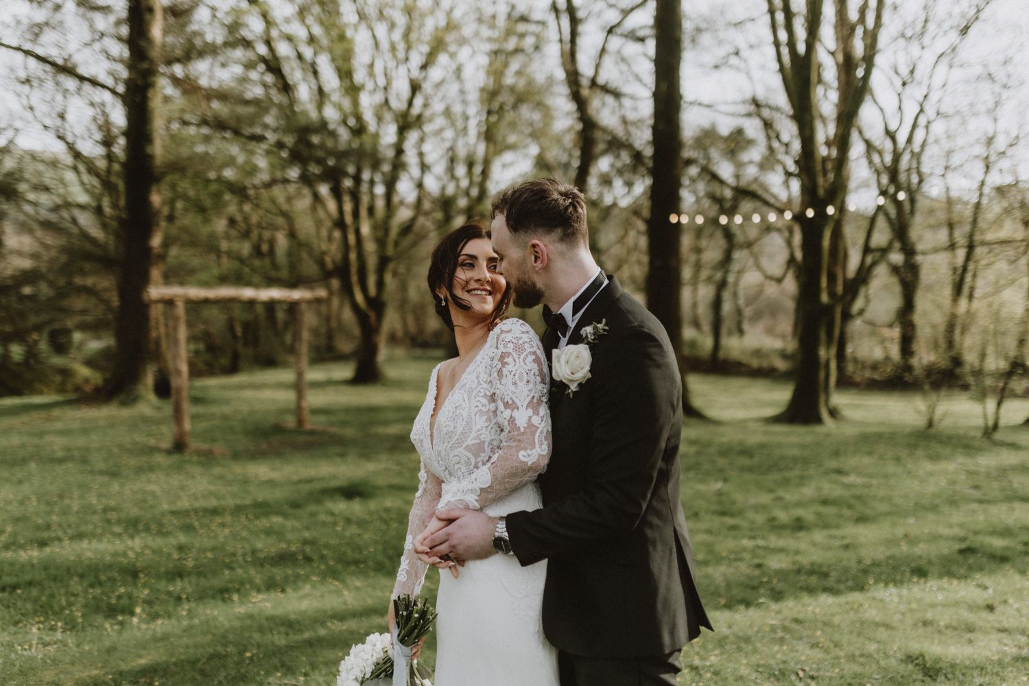 A couple in wedding attire share a romantic moment together on a green lawn surrounded by trees with string lights overhead.