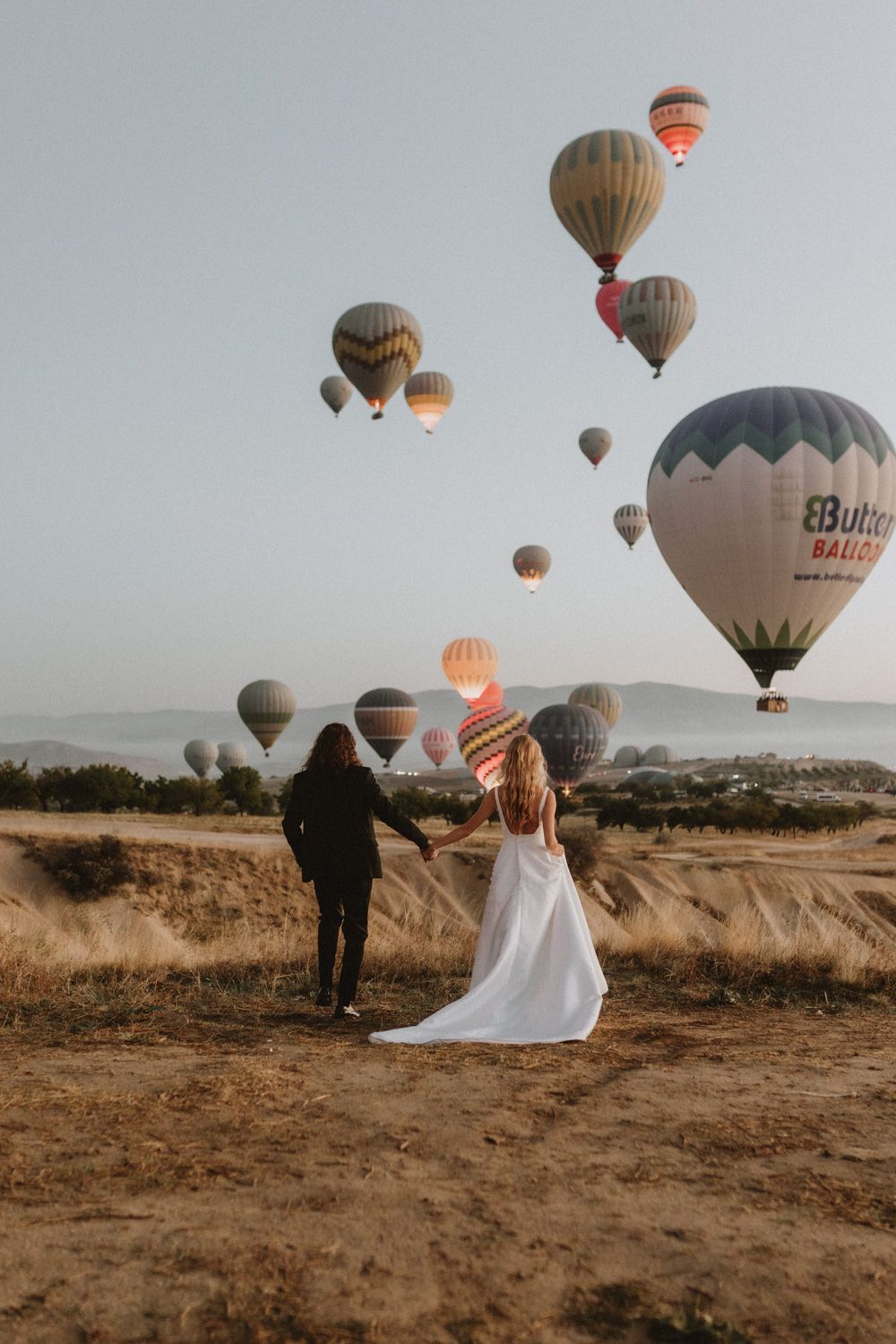 Hot air balloons float in the sky above a couple in formal attire on a desert hill during sunset.