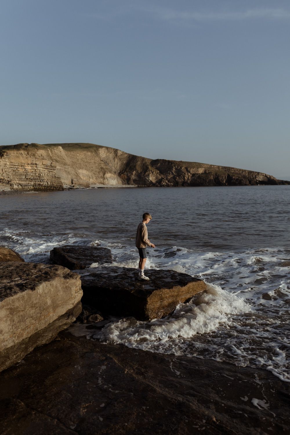 Person standing on rocky outcrop with dramatic coastal cliffs and crashing waves.