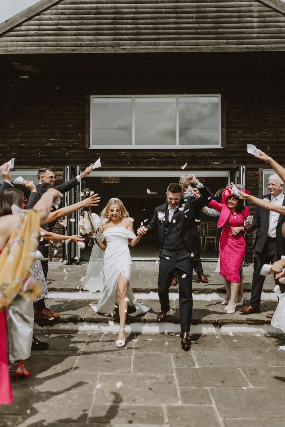 Wedding guests throwing confetti at celebrating couple outside rustic venue.