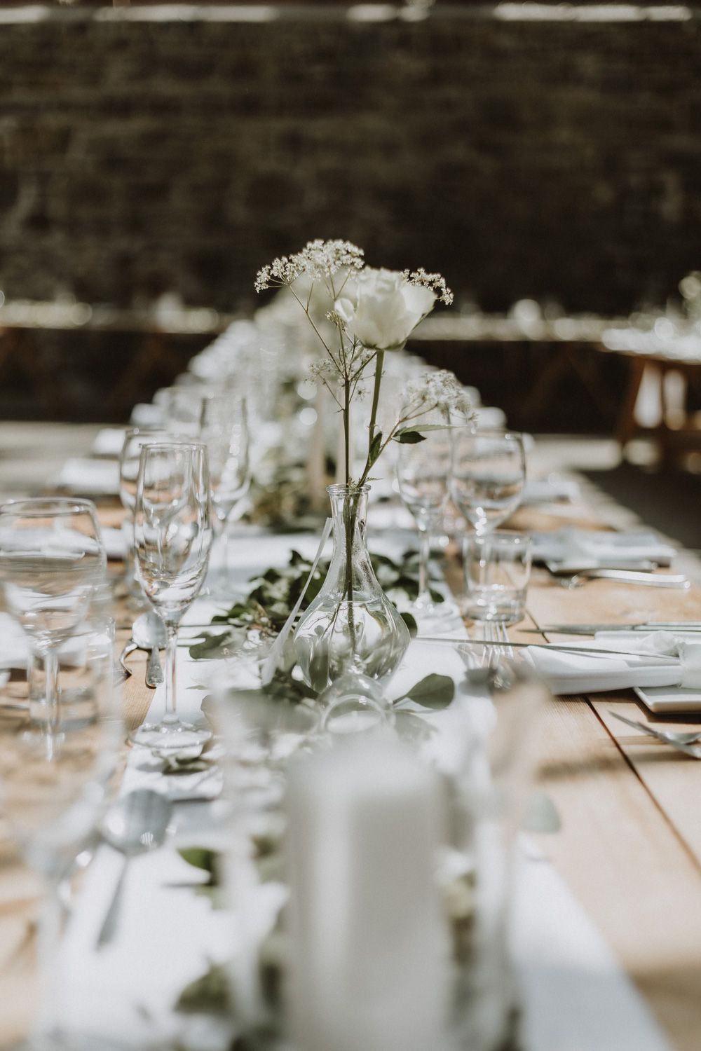 Elegant wedding table setting with delicate white floral centerpieces and wine glasses on rustic wooden table.